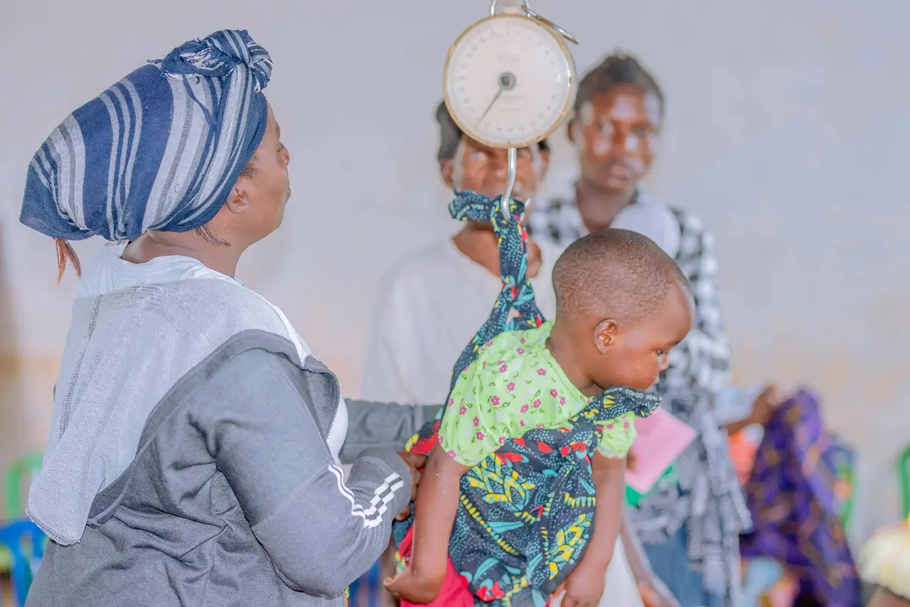 A health worker weighing a toddler in a sling scale while the mother looks on